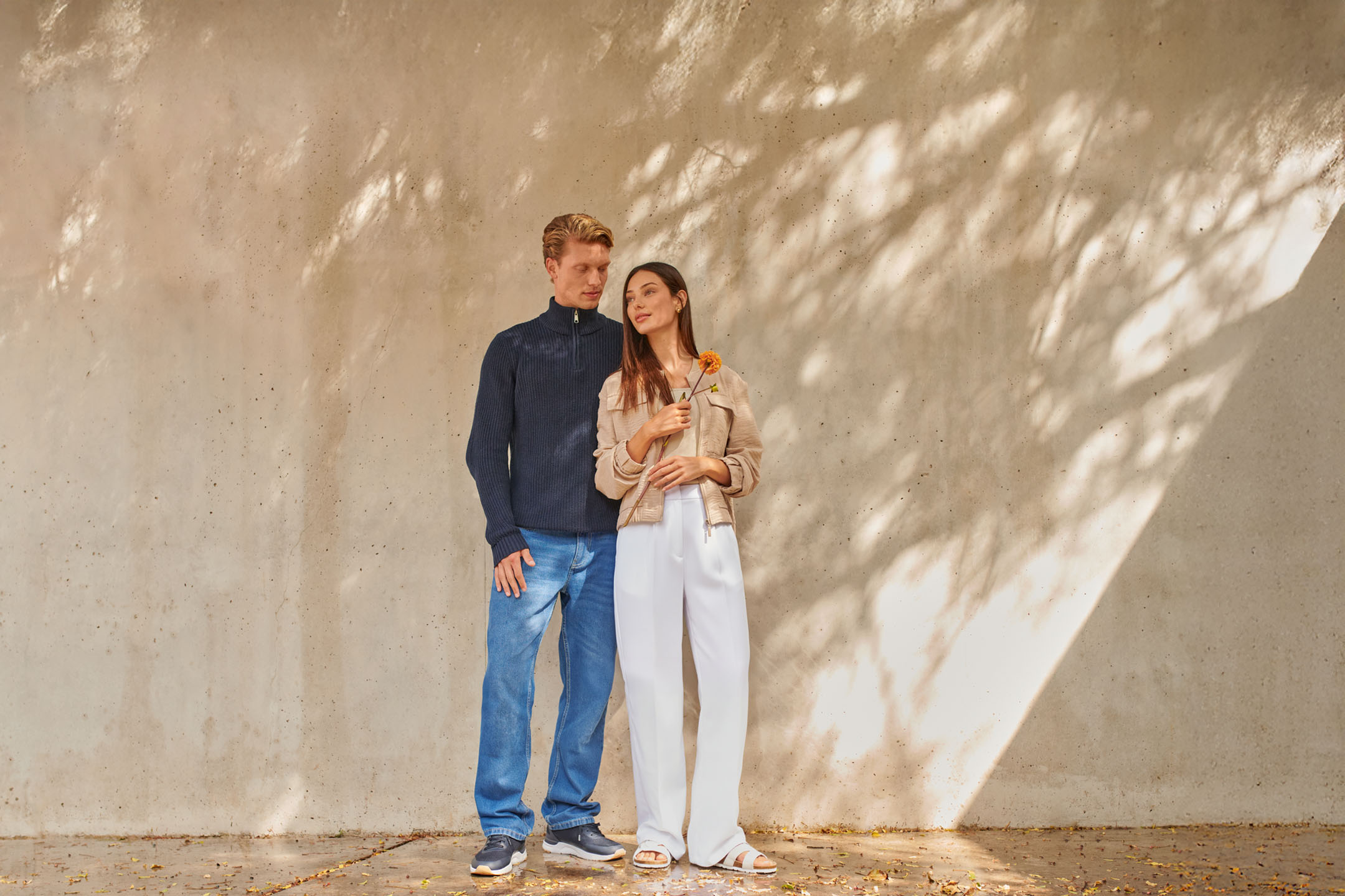 Homme en jean bleu et pull bleu foncé, femme en pantalon blanc et veste beige avec une fleur.