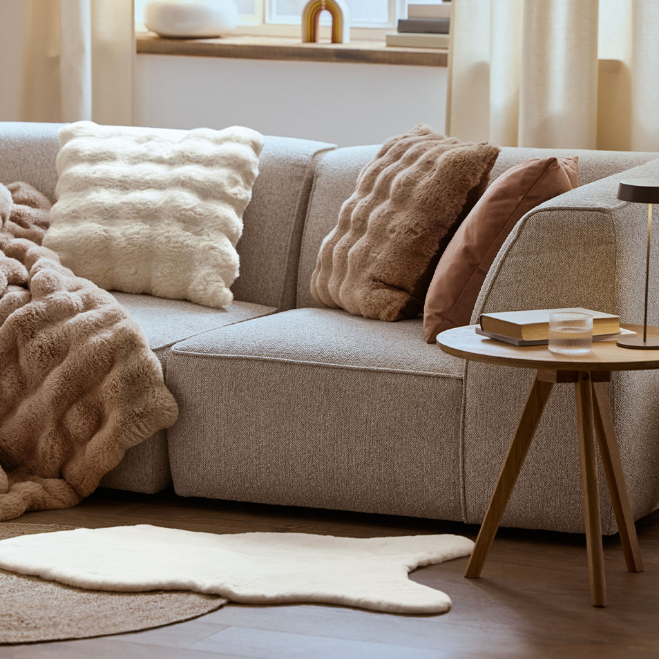 Un canapé modulaire gris avec des coussins et une couverture moelleux, à côté d'une table d'appoint en bois.