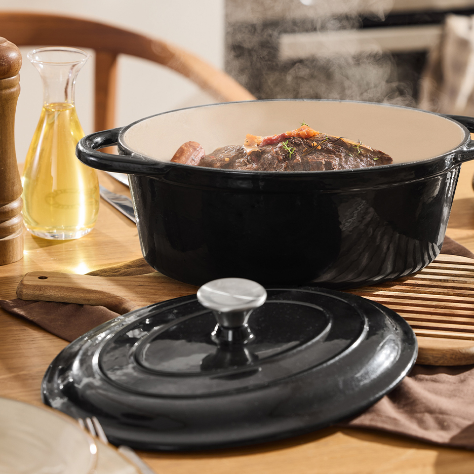 Cocotte ovale noire en fonte avec plat de viande fumant sur une table en bois.