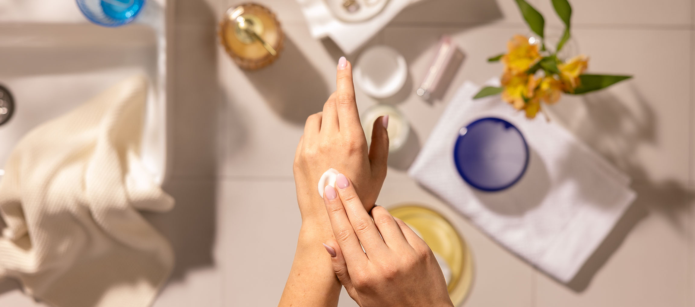 Mains féminines appliquant de la crème pour les mains, entourées de cosmétiques et serviettes dans une salle de bain.
