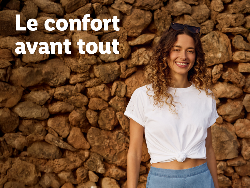 Une femme souriante en t-shirt blanc et pantalon bleu devant un mur de pierres, avec le texte 'Le confort avant tout'.