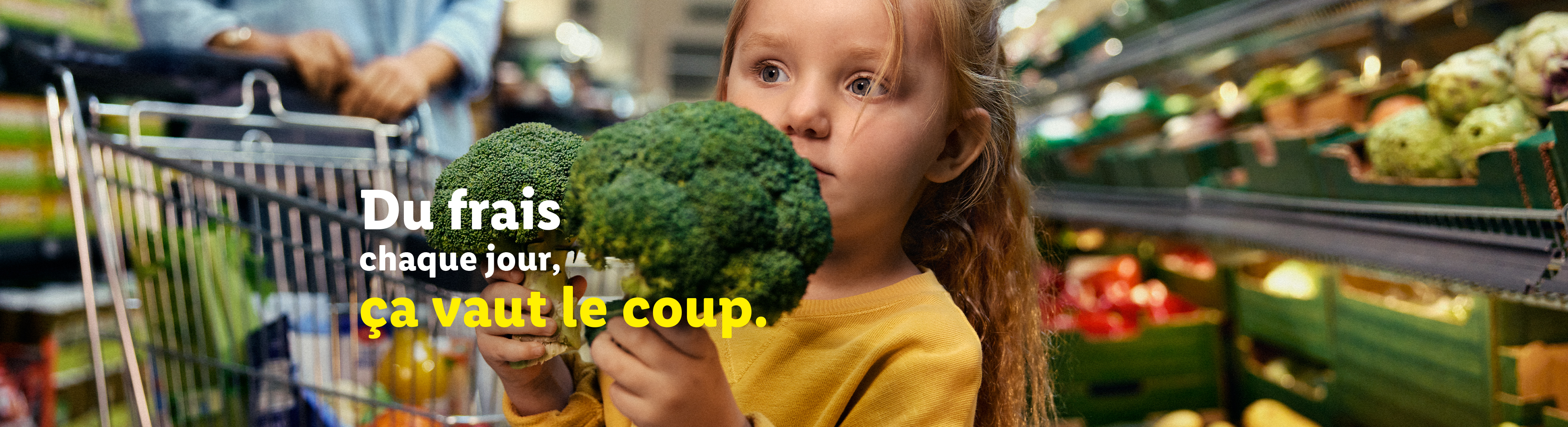 Une petite fille tient du brocoli dans une épicerie, avec un chariot en arrière-plan.