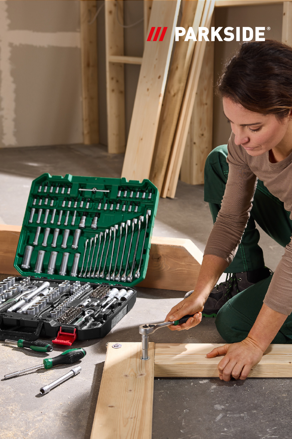 Une femme assemble des planches de bois avec une clé à cliquet, un grand coffret d'outils ouvert à côté.