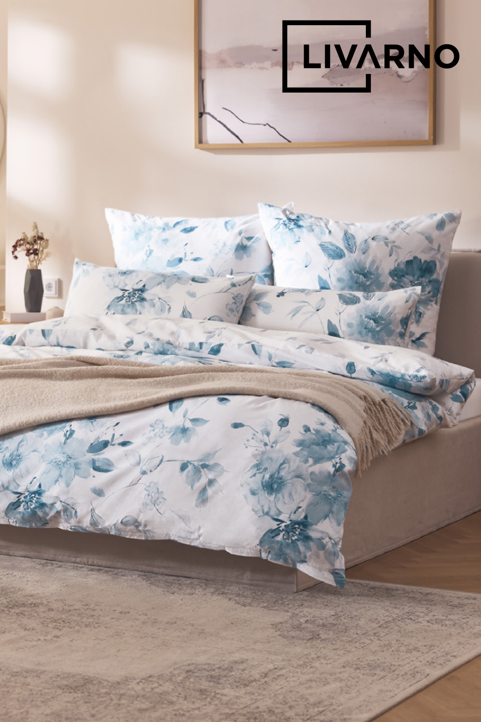 Parure de lit blanche à motifs floraux bleus sur un lit, avec un plaid beige et un tableau abstrait.