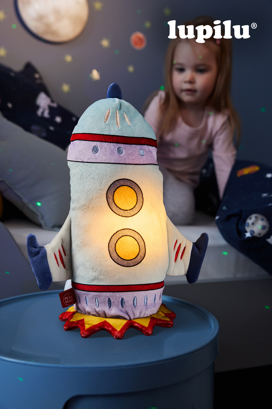 Lampe de nuit en forme de fusée en peluche avec une petite fille en arrière-plan.
