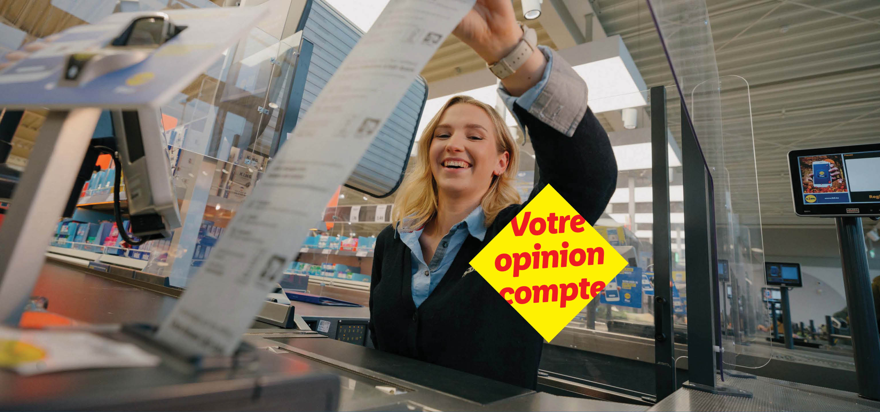 Une caissière souriante tient un long reçu, avec un panneau jaune 'Votre opinion compte'.