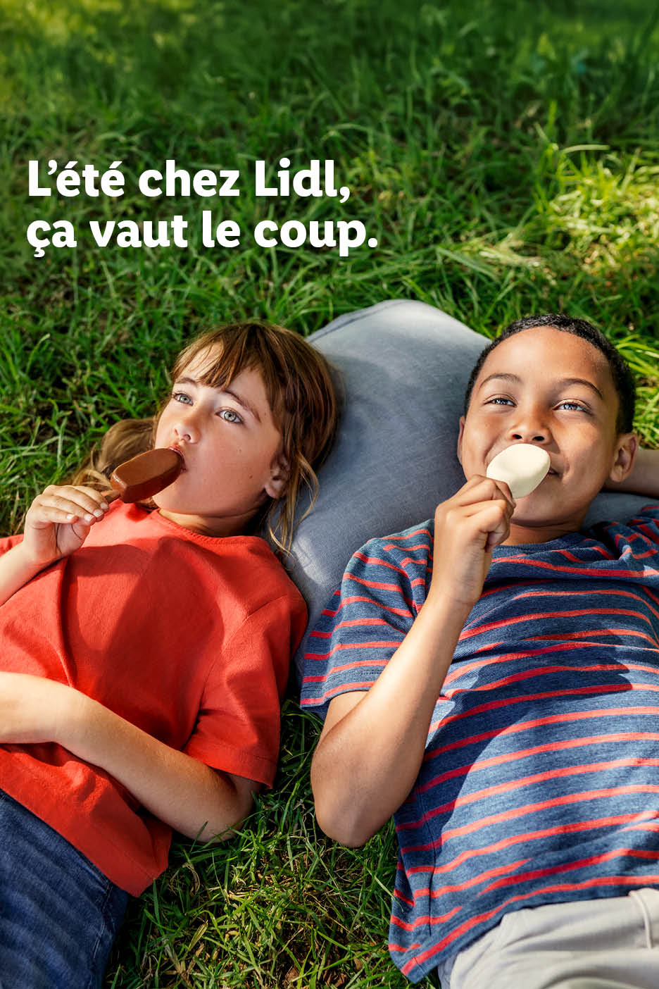 Deux enfants allongés dans l'herbe, mangeant des bâtonnets glacés. Texte: L'été chez Lidl, ça vaut le coup.