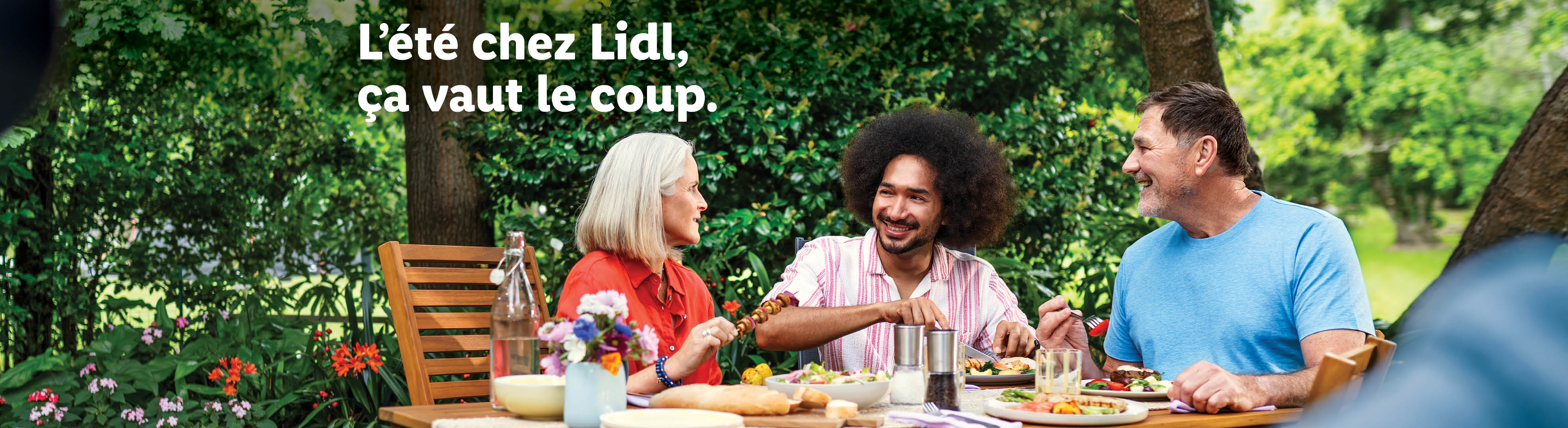 Des amis souriants partagent un repas d'été en plein air, avec des brochettes et de la salade. Texte : L'été chez Lidl, ça vaut le coup.