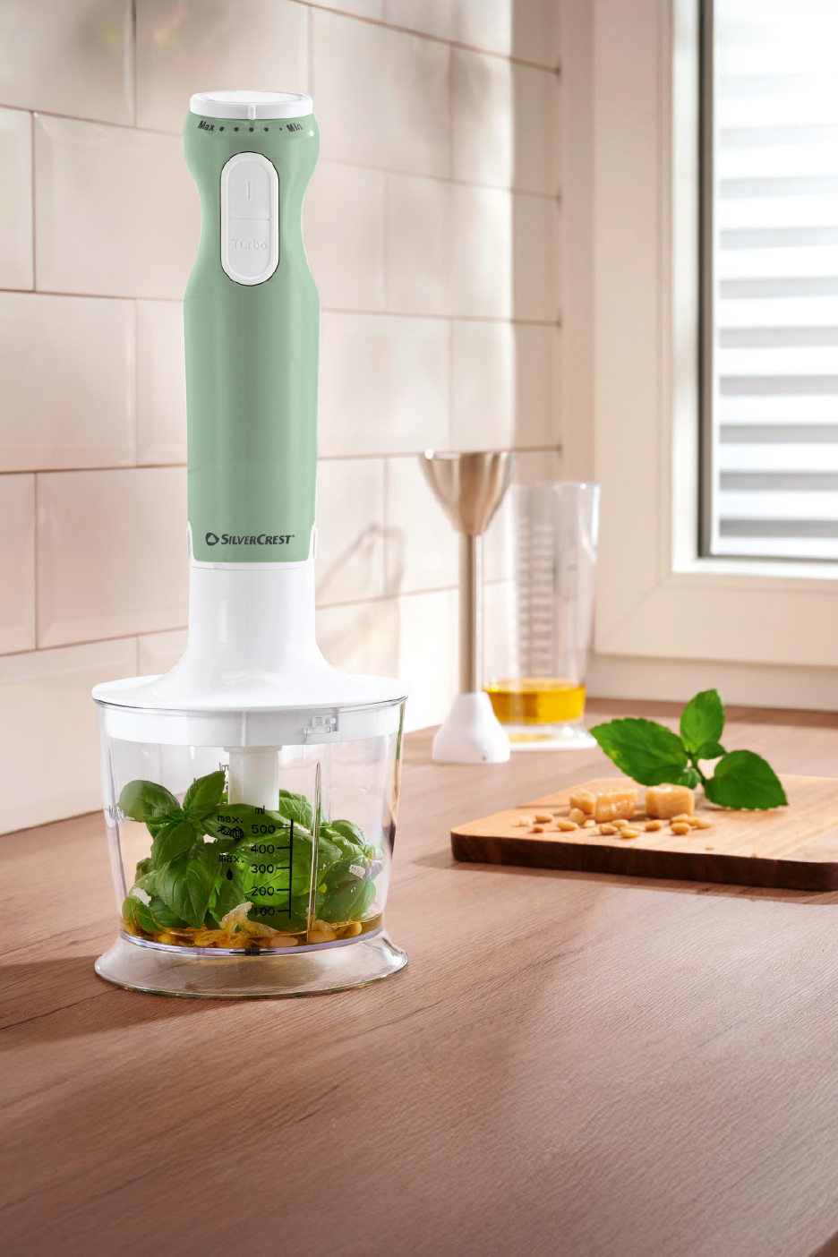 Mixeur plongeant vert et blanc préparant du pesto avec basilic, pignons et parmesan.