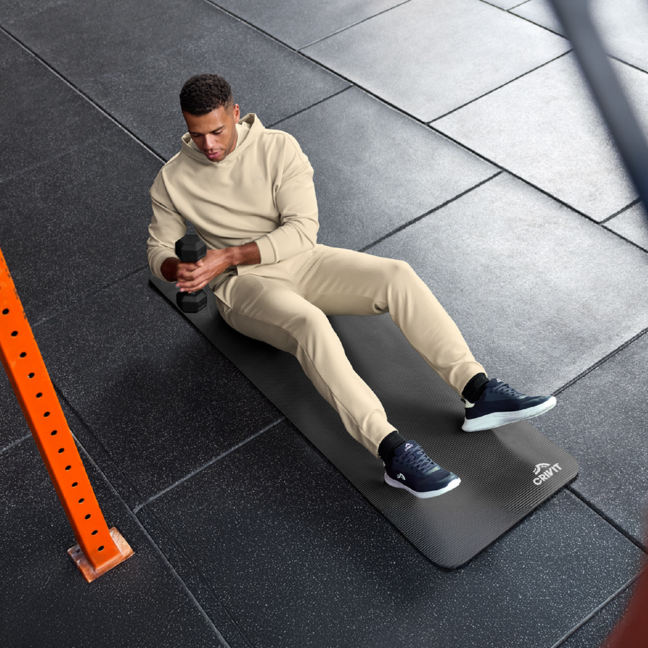 Un homme en survêtement beige fait des exercices avec un haltère sur un tapis de fitness.