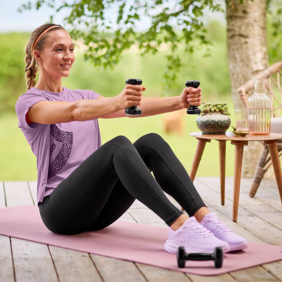 Femme en t-shirt violet et leggings noirs faisant de l'exercice avec des haltères sur un tapis de yoga.