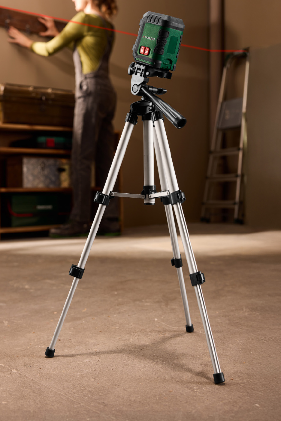 Niveau laser sur trépied projetant une ligne rouge, avec une personne travaillant en arrière-plan.