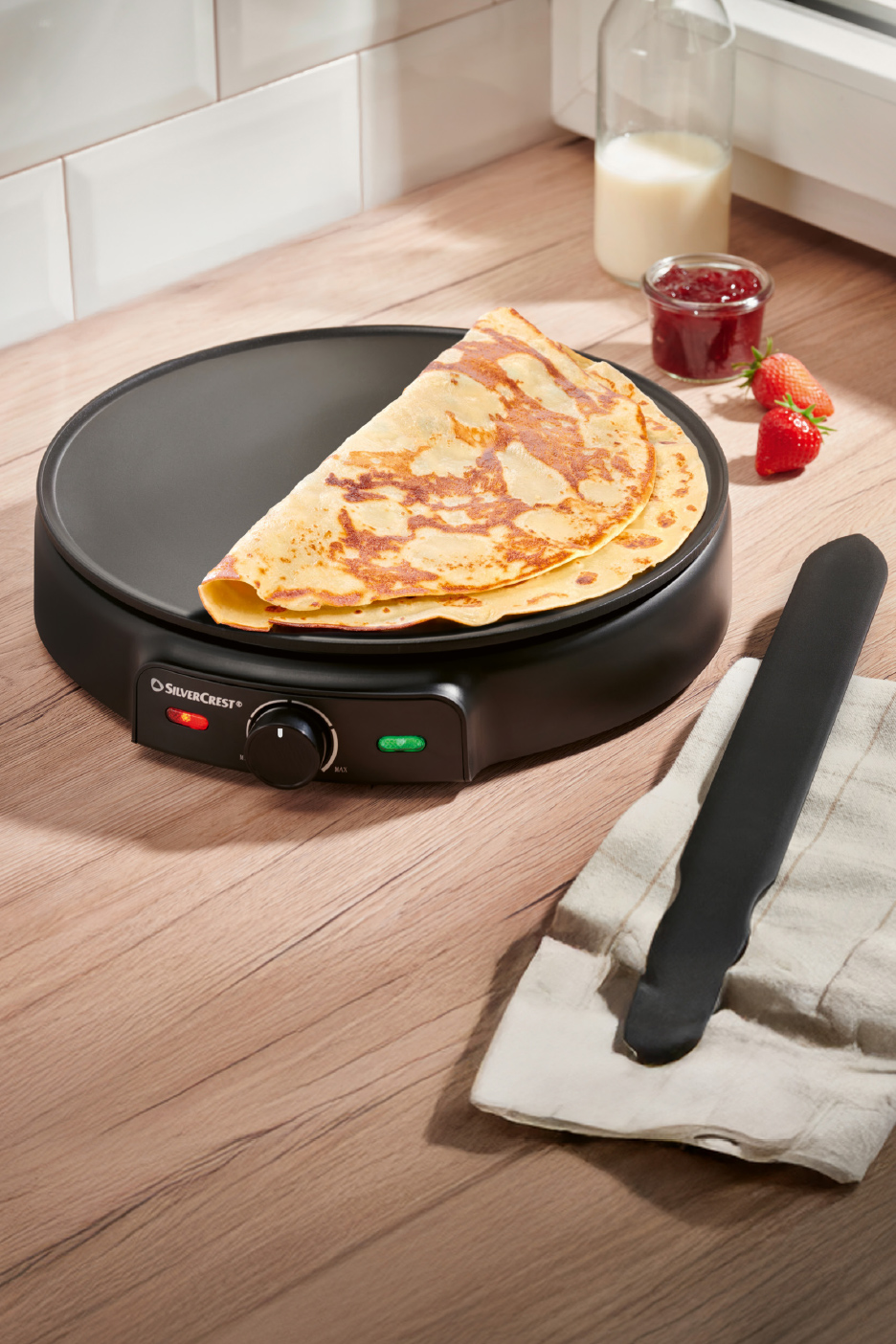 Crêpière électrique avec une crêpe, du lait, de la confiture et des fraises sur un comptoir en bois.