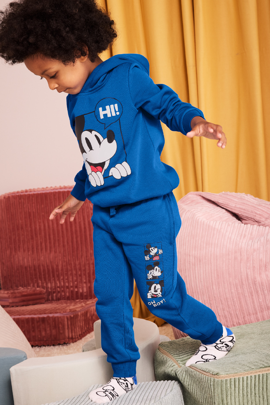 Un enfant en sweat à capuche et pantalon de survêtement bleus Mickey Mouse se tient sur des blocs moelleux.