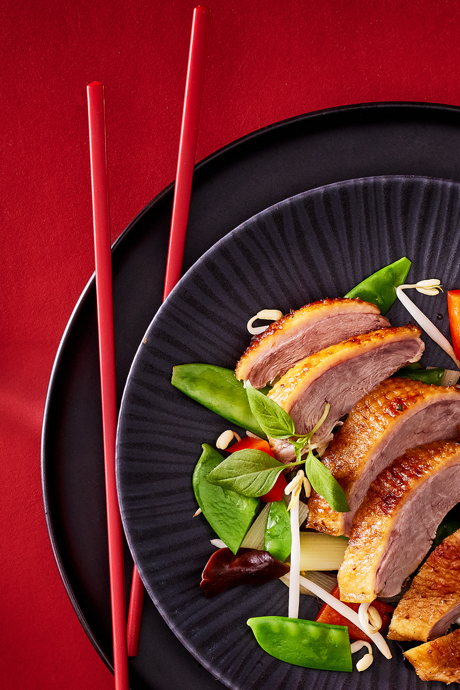 Canard laqué avec légumes et baguettes rouges sur fond rouge.