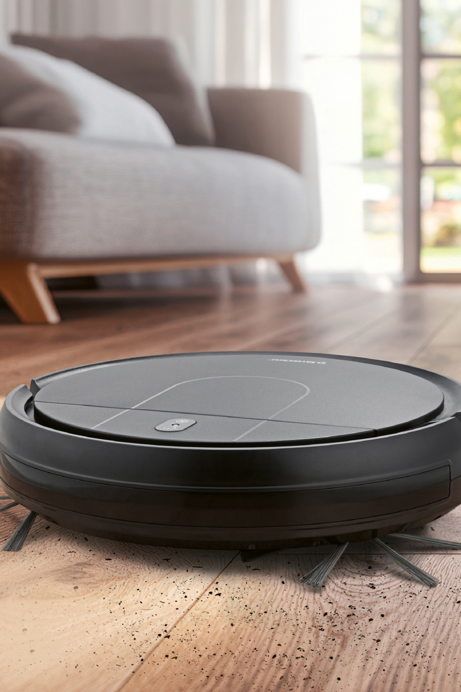 Aspirateur robot noir sur un sol en bois sale, avec un canapé en arrière-plan.