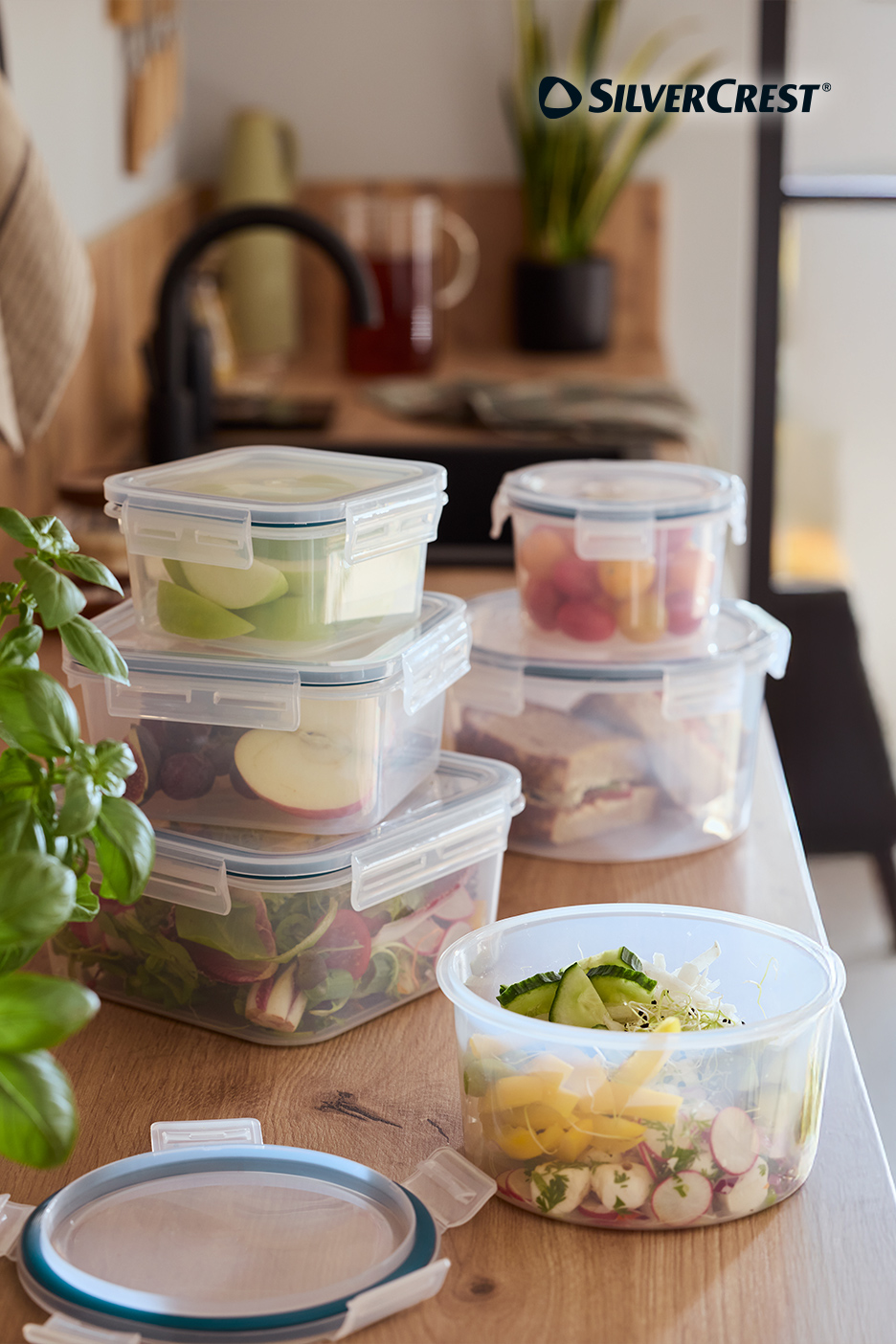 Récipients alimentaires transparents remplis de fruits, légumes et sandwichs sur un comptoir de cuisine en bois.