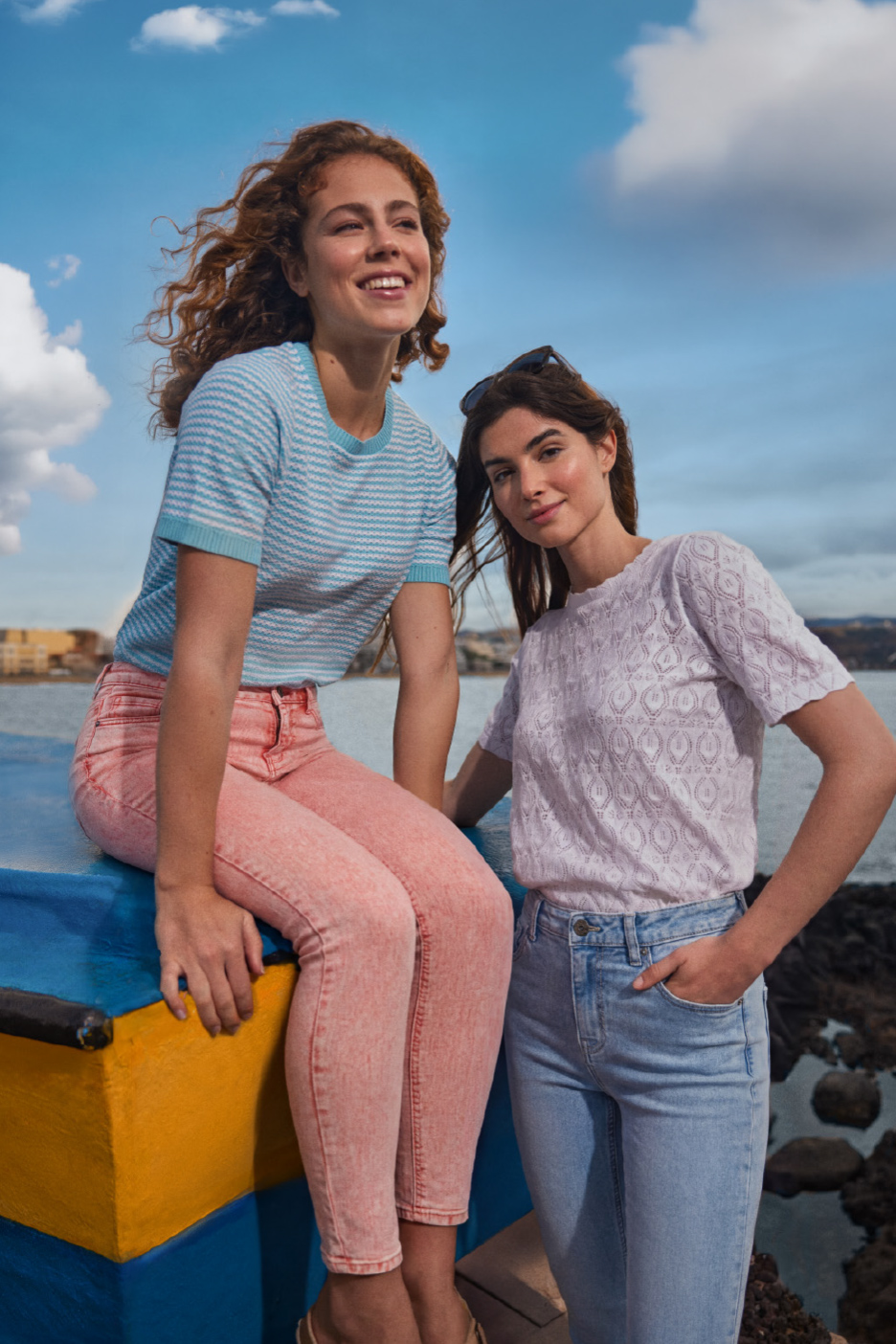 Deux femmes en jeans et hauts d'été, l'une assise, l'autre debout, près de l'eau.