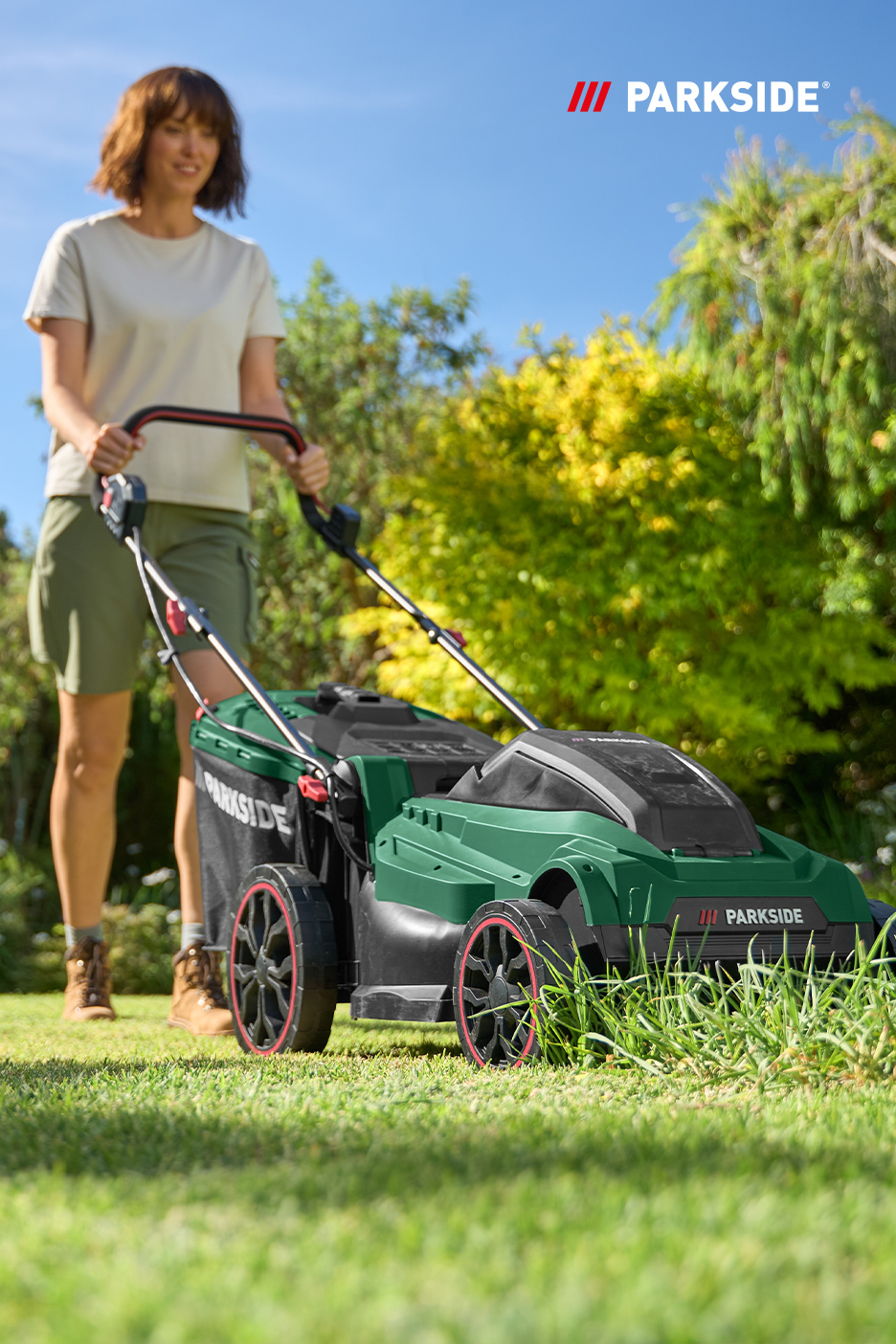 Une femme tond la pelouse avec une tondeuse à gazon verte dans un jardin ensoleillé.
