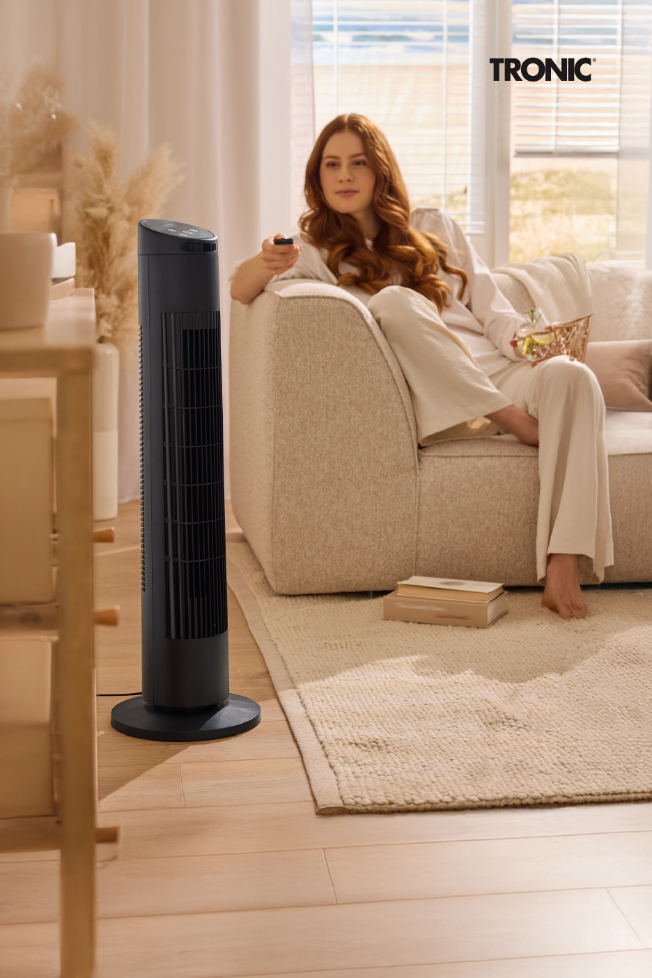 Une femme rousse se détend sur un canapé avec une télécommande, un ventilateur colonne noir à côté.