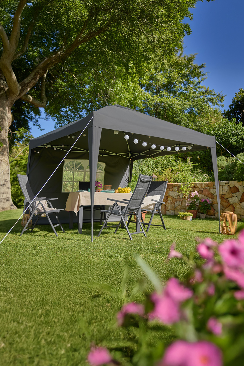 Tonnelle de jardin grise avec table et chaises, décorée de guirlandes lumineuses, sur une pelouse.
