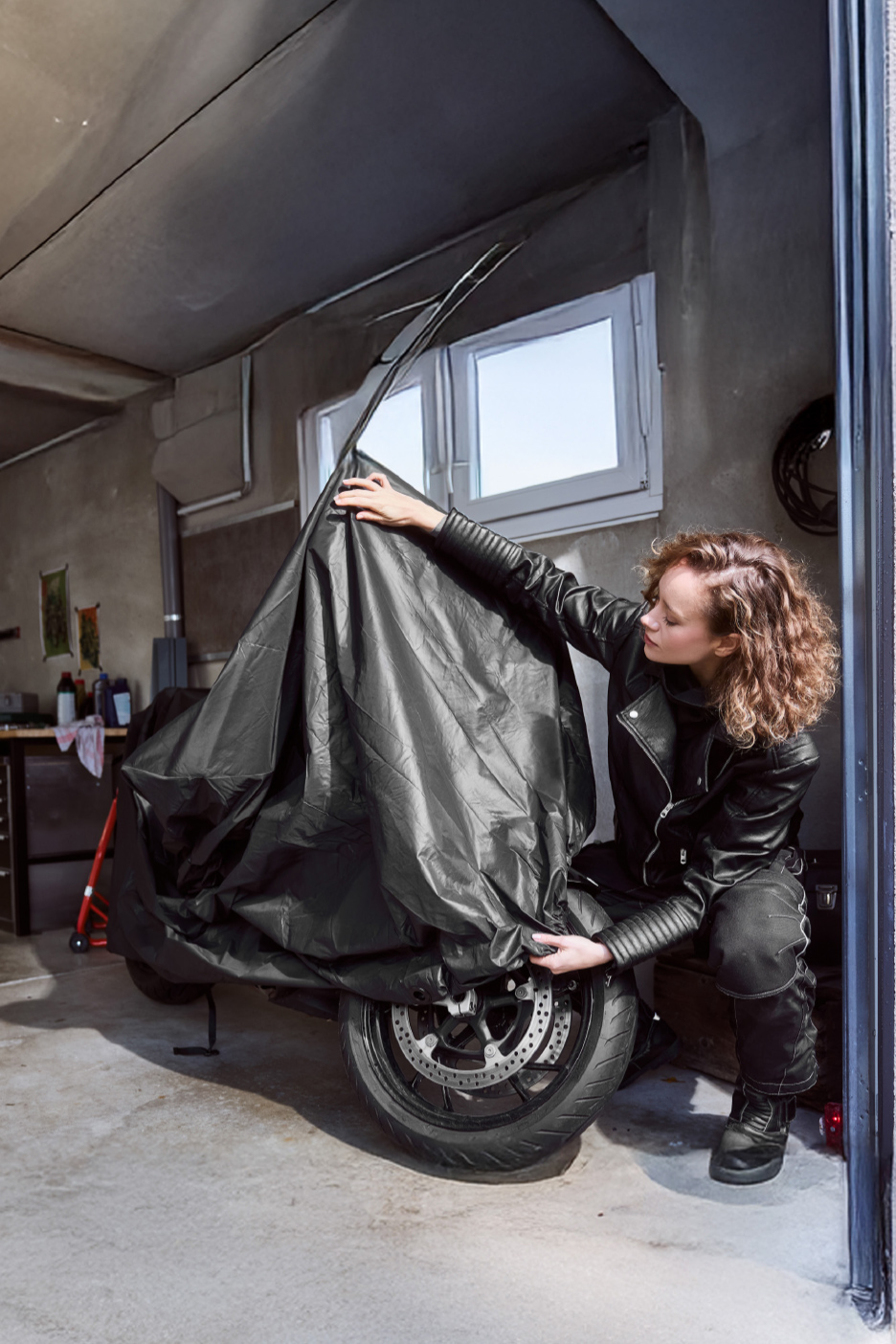Femme en veste de motard noire couvrant une moto avec une bâche de protection dans un garage.