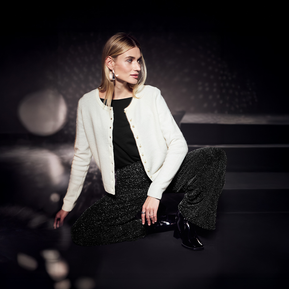 Femme en cardigan blanc, haut noir et pantalon scintillant noir, accroupie.