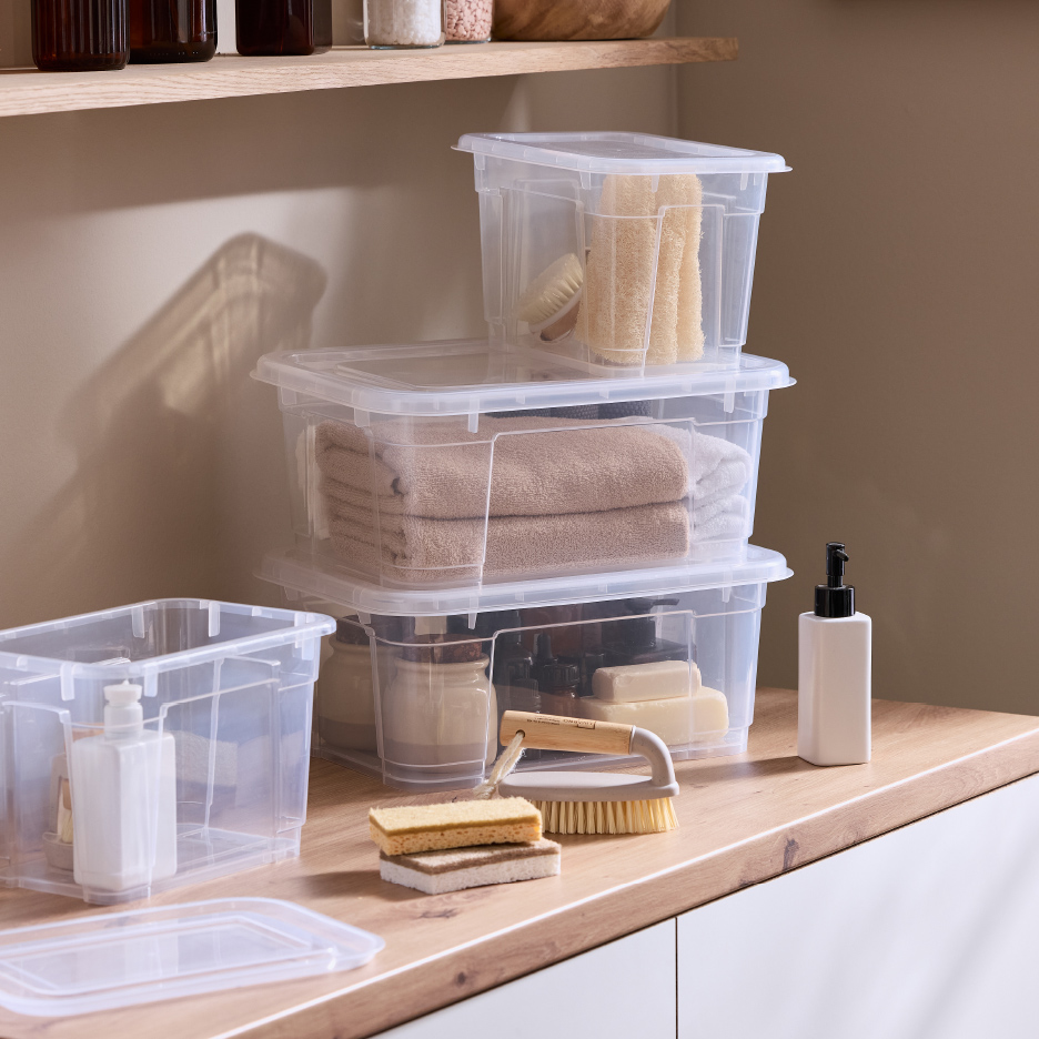 Boîtes de rangement transparentes empilées avec serviettes, éponges et brosses sur un comptoir en bois.