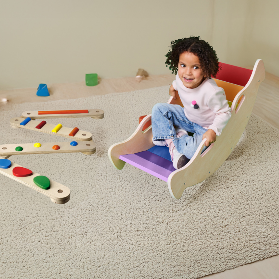 Enfant souriant sur un cheval à bascule arc-en-ciel, avec des jeux d'équilibre en bois sur un tapis.