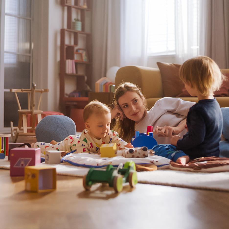Mère et ses deux enfants jouent avec des jouets en bois et des blocs colorés sur un tapis.