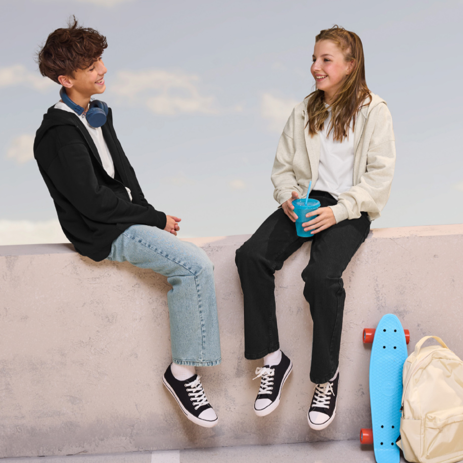 Deux adolescents souriants, vêtus de sweats à capuche et de jeans, assis sur un mur avec un skateboard et un sac à dos.