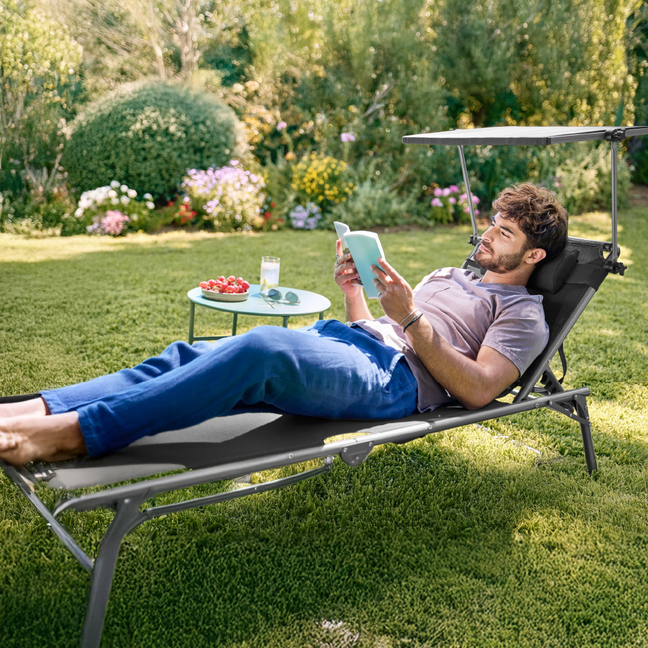 Homme lisant sur un transat de jardin avec pare-soleil, à côté d'une table basse avec boisson et fruits.