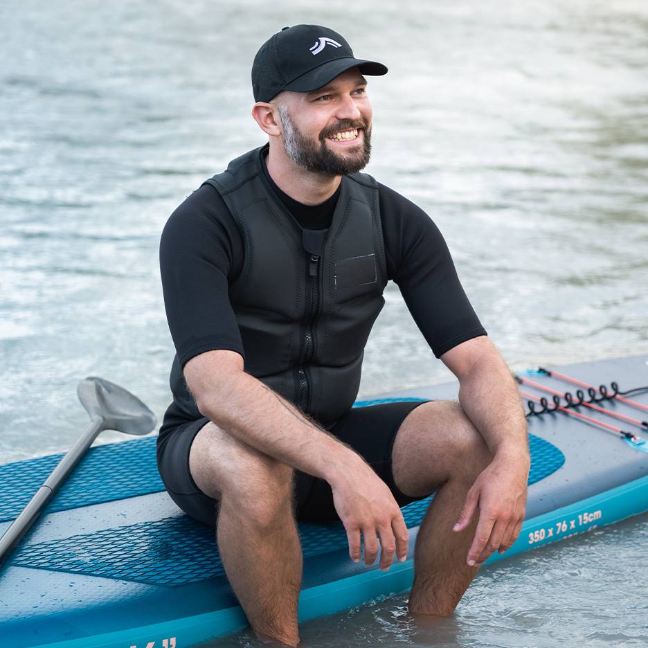 Homme souriant avec gilet de sauvetage et casquette, assis sur une planche de paddleboard dans l'eau.