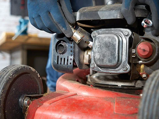 Mains gantées remplaçant la bougie d'allumage d'une tondeuse rouge.