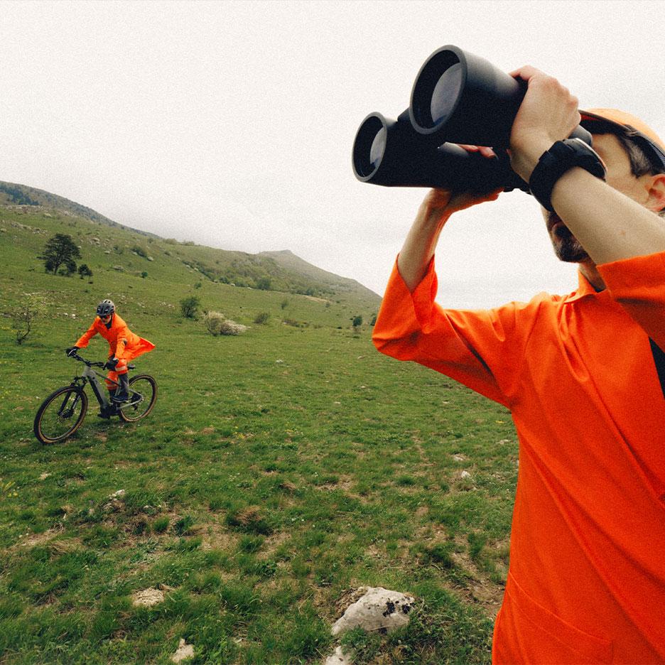 Homme avec des jumelles et cycliste en tenue de sport orange dans un paysage montagneux.