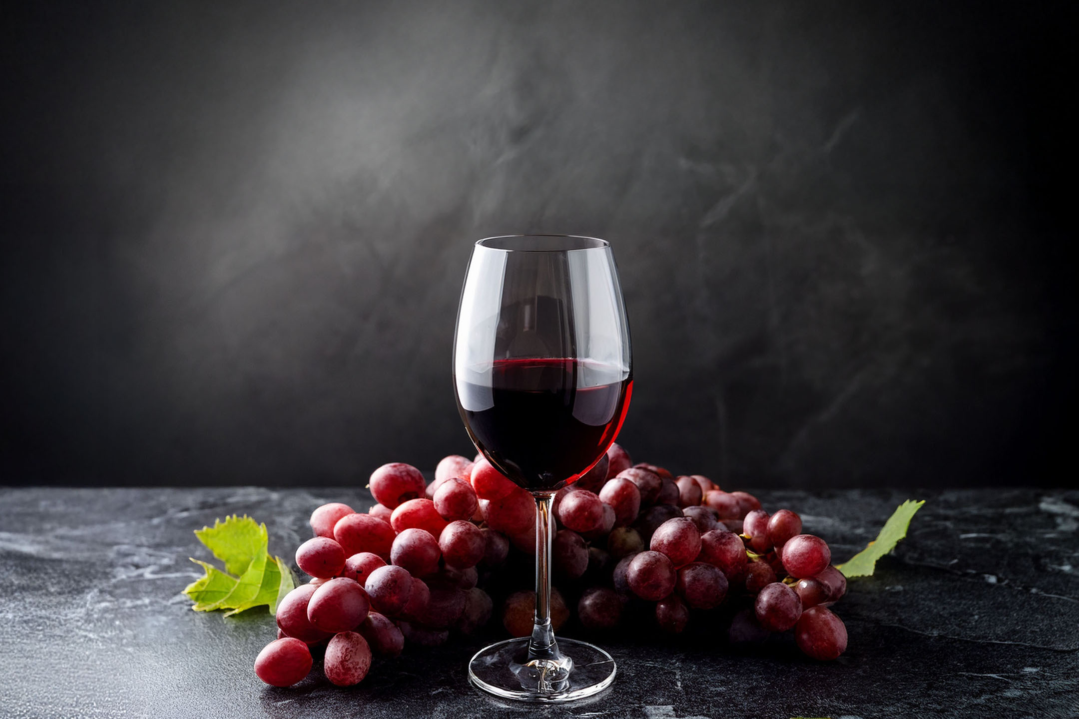 Un verre de vin rouge et une grappe de raisins rouges sur fond sombre.