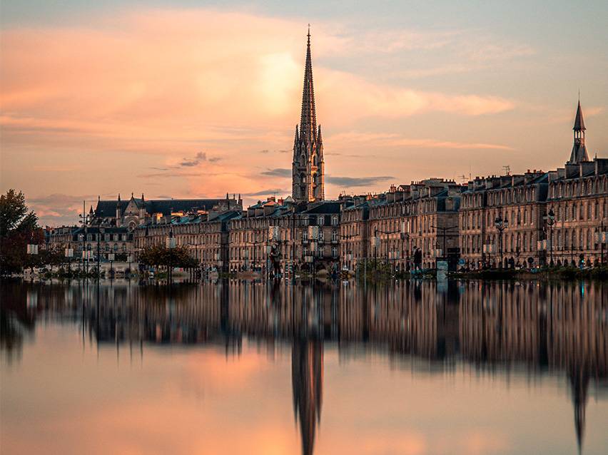 Paysage urbain de Bordeaux au coucher du soleil avec une église et son reflet dans l'eau.
