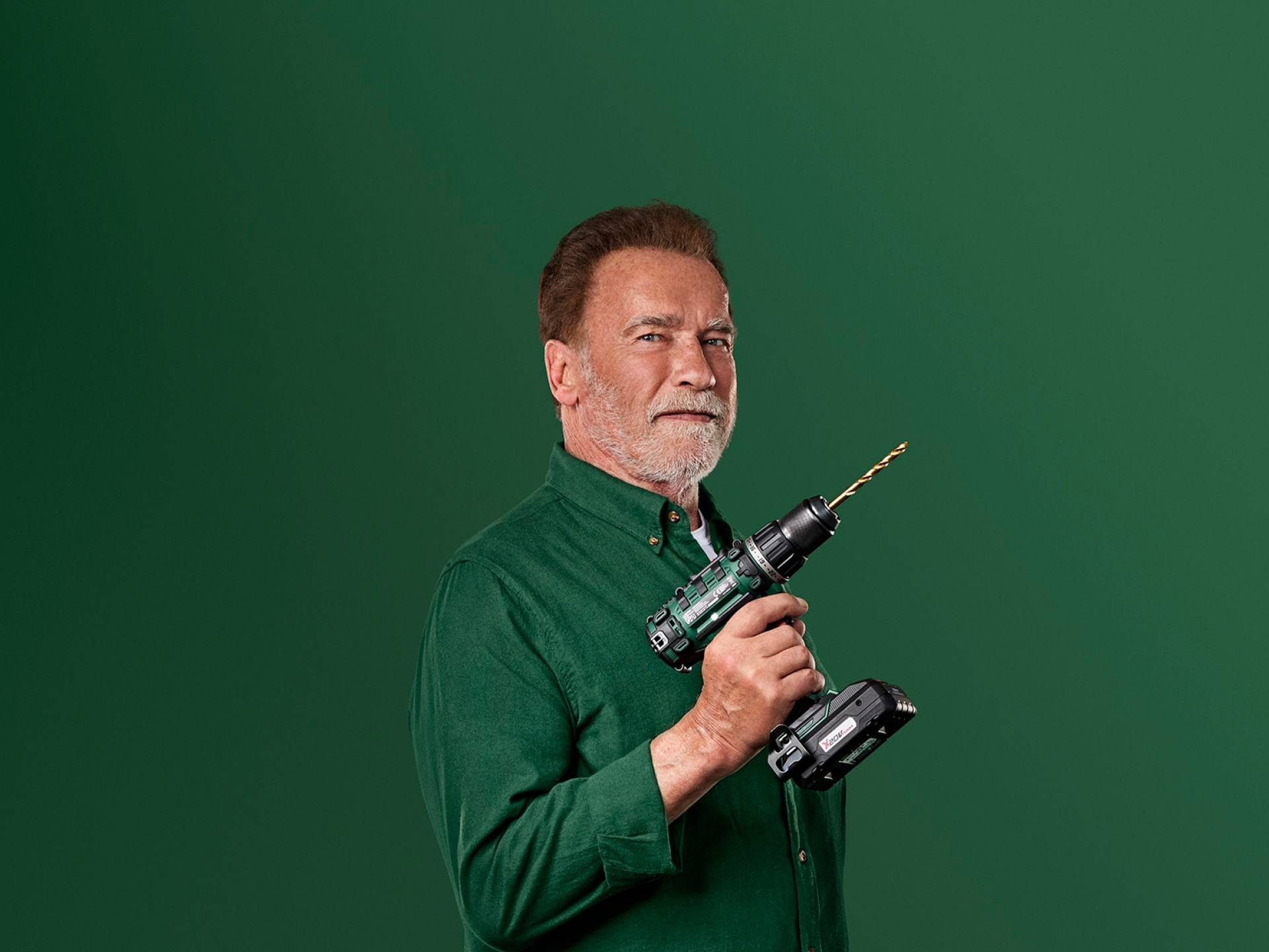 Arnold Schwarzenegger tient une perceuse électrique verte sur un fond vert.