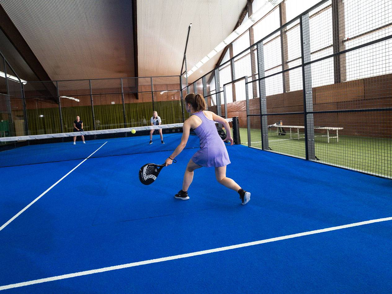 Femme jouant au padel sur un court bleu, avec d'autres joueurs en arrière-plan.