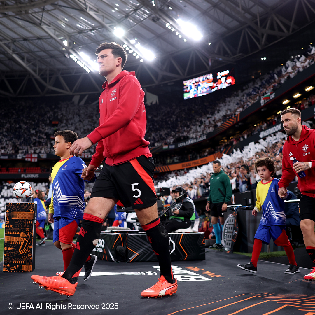 Des joueurs de football en maillots rouges et shorts noirs marchent sur un terrain avec des enfants, vêtus de tenues de sport.