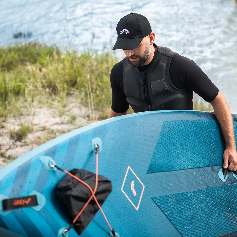 Homme en gilet de sauvetage et casquette tenant une planche de paddleboard bleue près de l'eau.