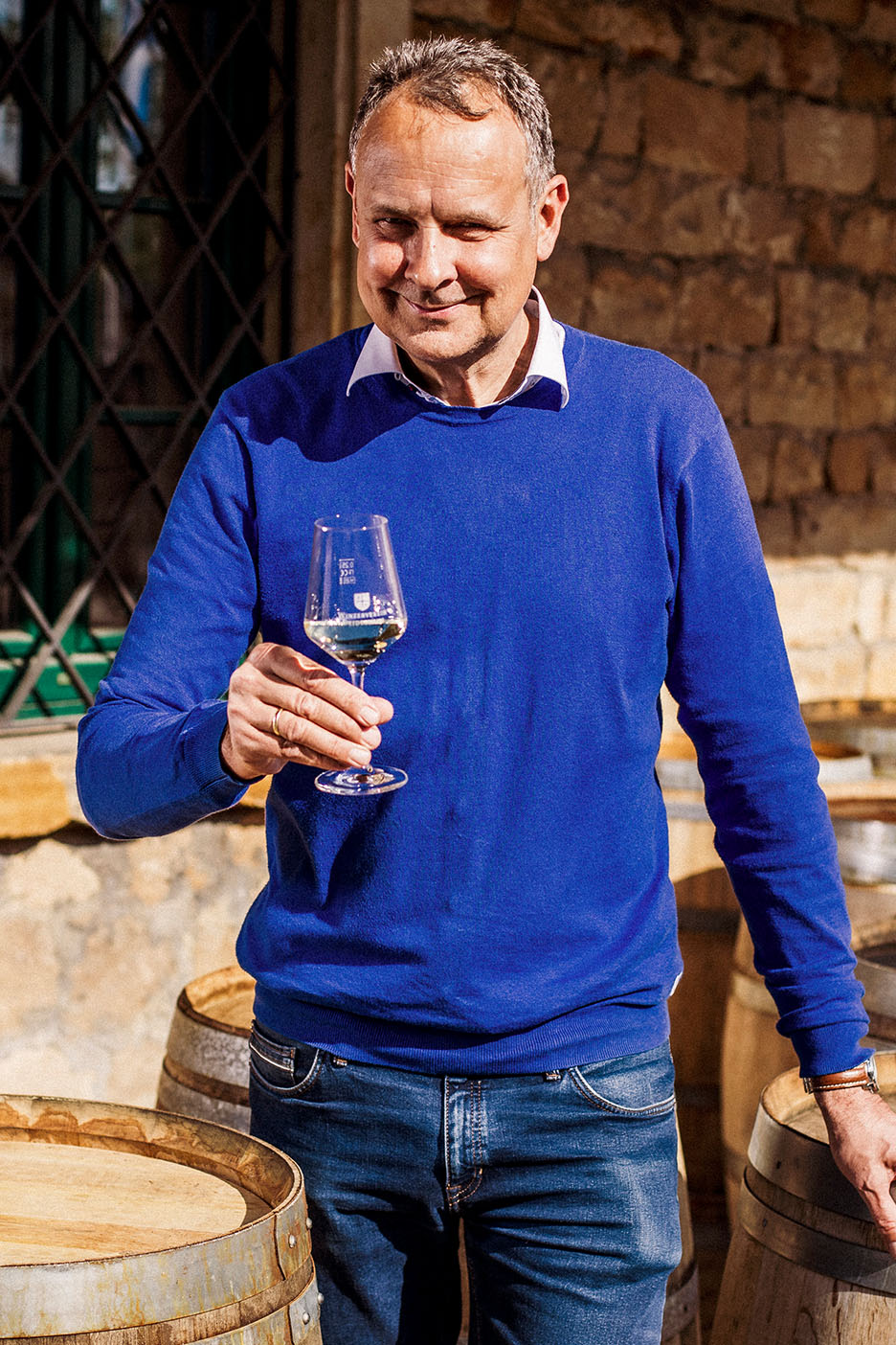 Homme souriant en pull bleu et jean, tenant un verre de vin blanc parmi des tonneaux en bois.