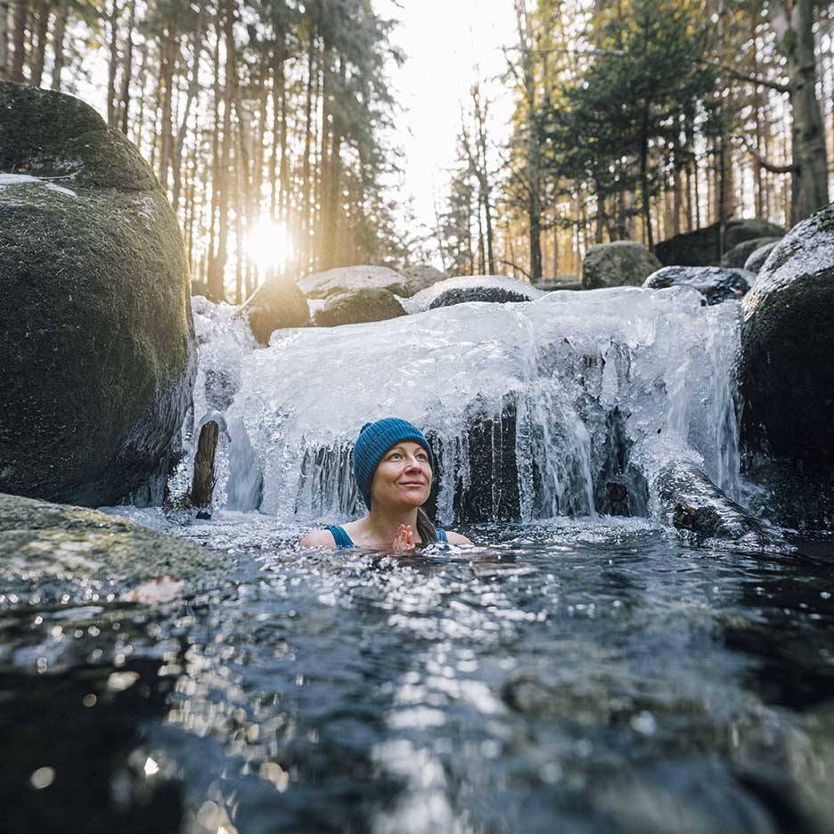 Femme en bonnet et maillot de bain bleus profitant d'un bain froid dans une rivière gelée.