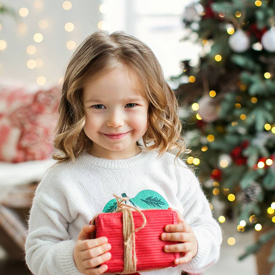 Fille souriante avec un cadeau de Noël, avec un sapin de Noël illuminé en arrière-plan.