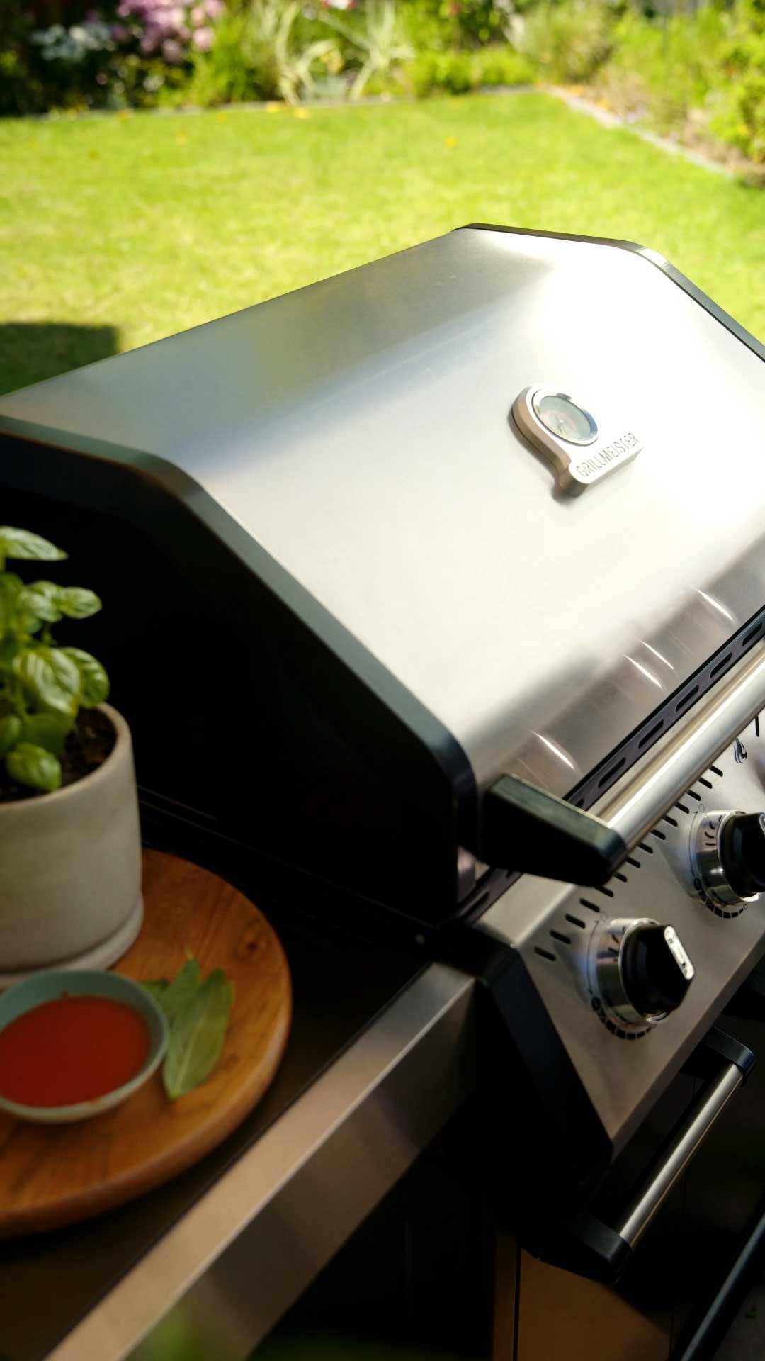 Barbecue à gaz en acier inoxydable avec thermomètre, à côté d'un pot de basilic et de sauce.