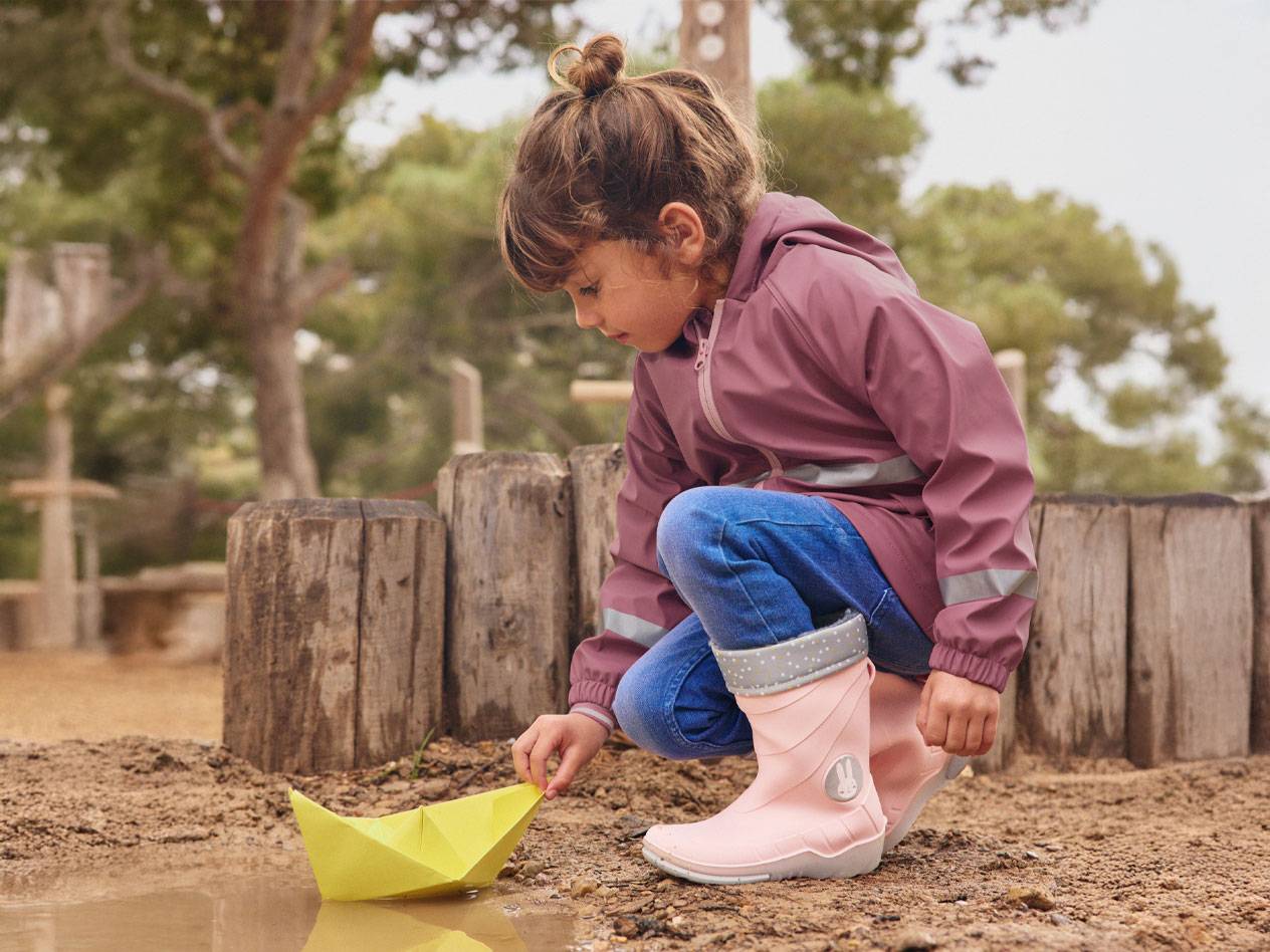 Fillette en bottes de pluie roses et veste, jouant avec un bateau en papier dans une flaque.