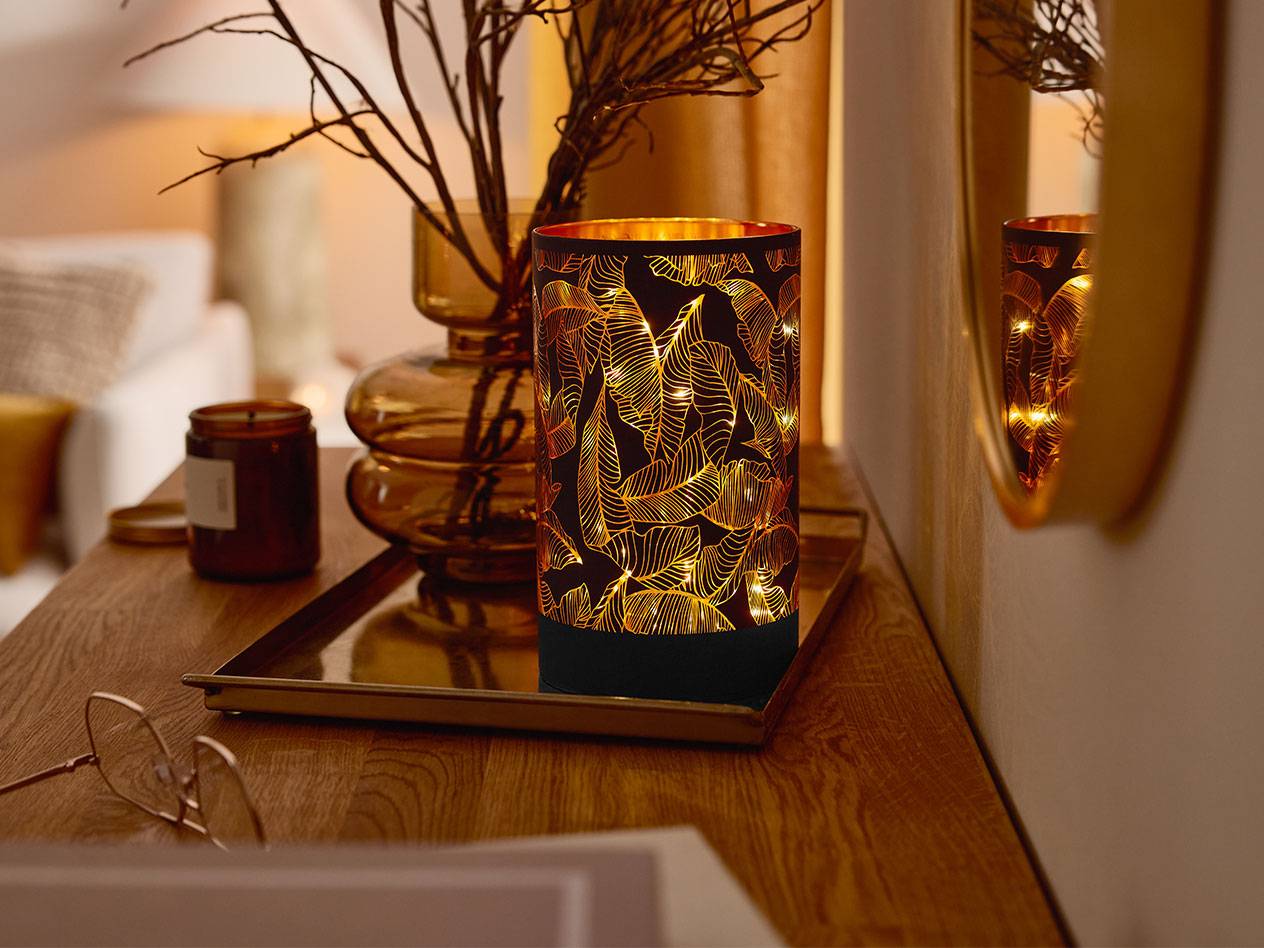 Une lanterne LED décorative avec un motif de feuilles, un vase avec des branches et une bougie sur une table en bois.
