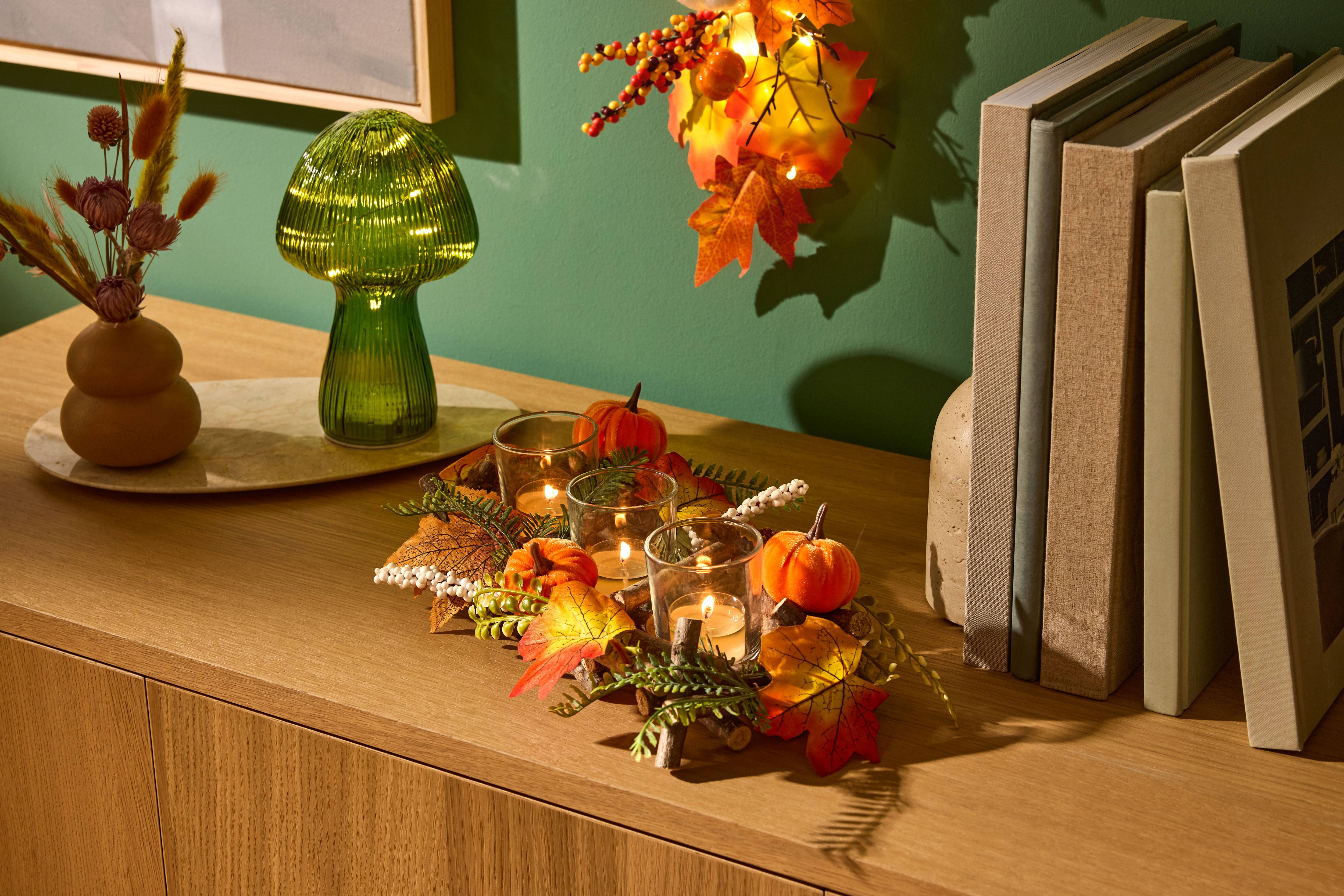 Décoration de table automnale avec citrouilles, bougies et feuilles, à côté d'une lampe champignon verte et de livres.
