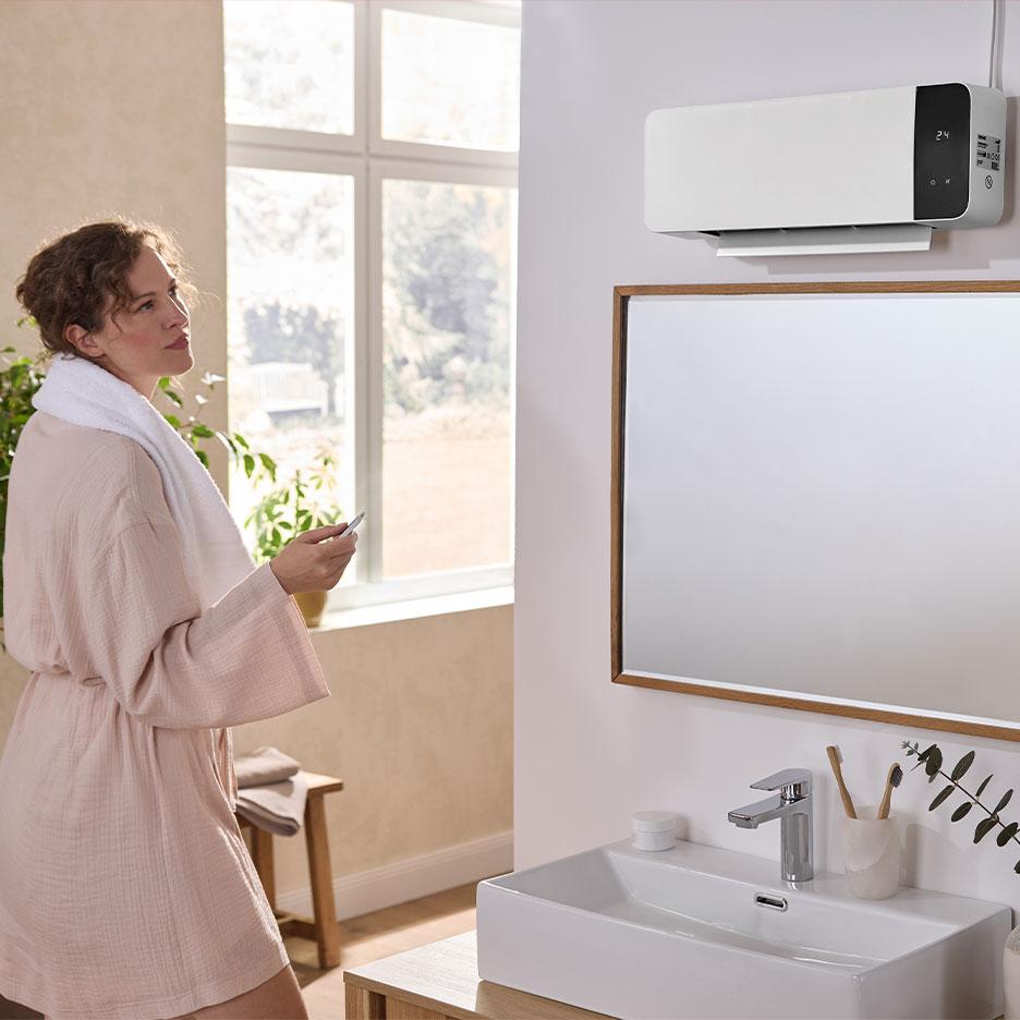 Une femme en peignoir utilise une télécommande pour régler un radiateur de salle de bain mural.