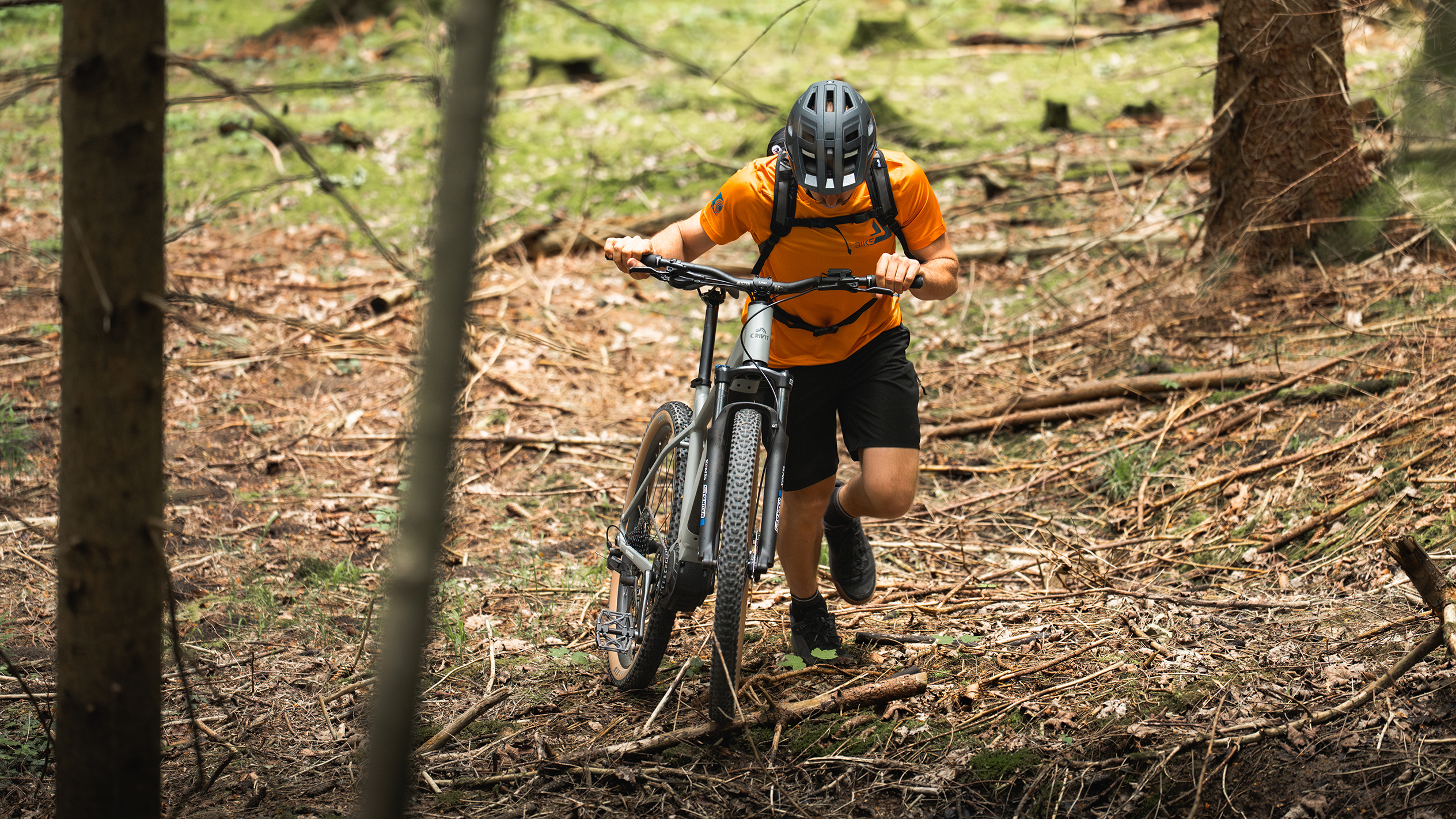 Un cycliste en maillot orange pousse un vélo électrique de montagne dans une forêt.